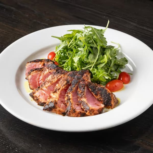 Seared tuna slices with a dark crust, pink center, green arugula, and cherry tomatoes on a white plate.