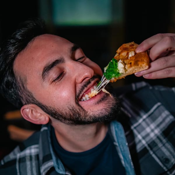 A man with a beard happily takes a bite of a pizza slice with stringy cheese and broccoli.